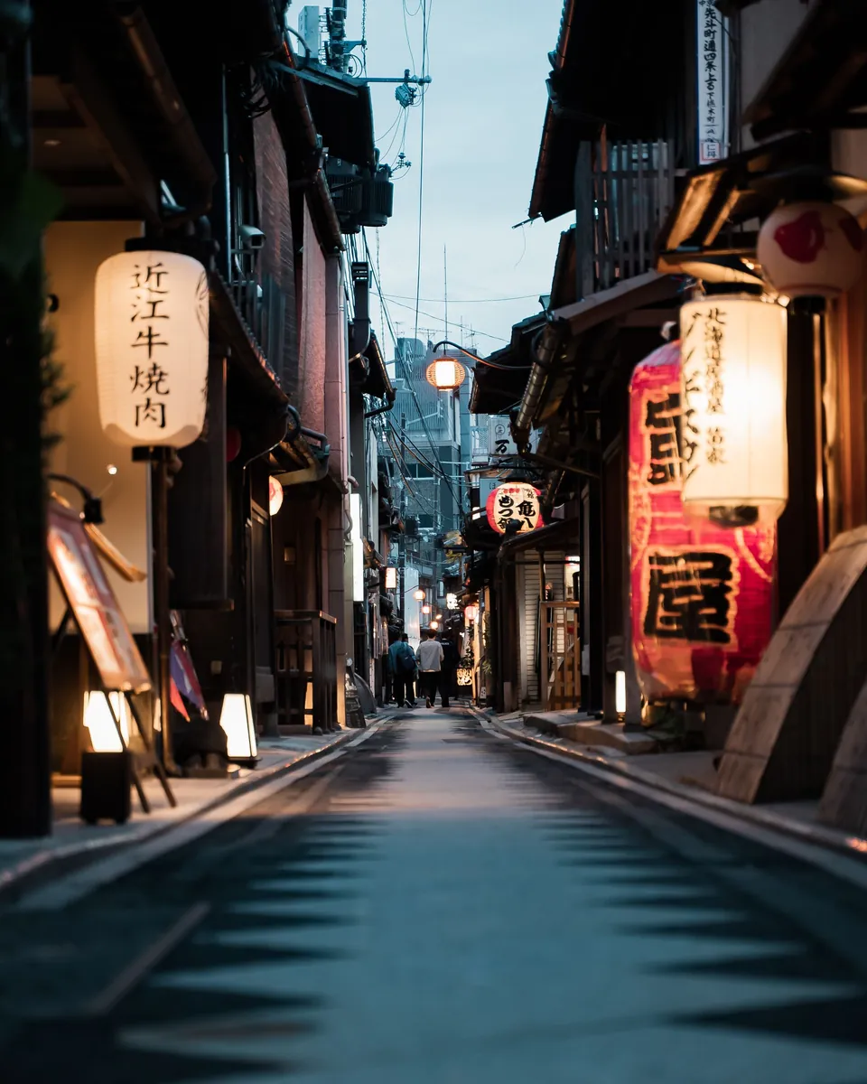 A narrow lane in Kyoto at dusk, lined with wooden shopfronts and paper lanterns